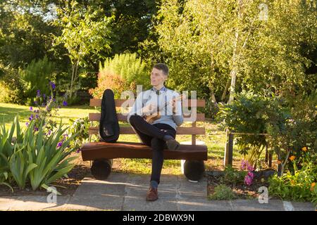 Young man sitting on bench in the park and playing ukulele. Stock Photo