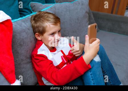 Handsome kid boy in Santa Claus hat looks at smartphone screen, shows thumb up.Talk with family,closeup image. Stock Photo