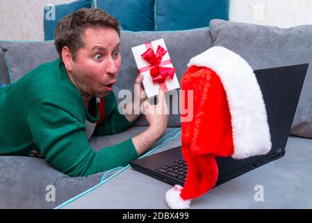 Surprised Male man virtually exchanging Christmas gifts and having video call over a laptop at home on the couch. Stock Photo