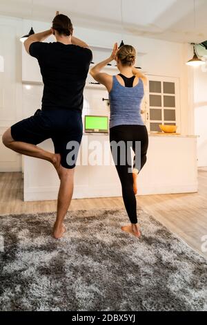 White caucasian fit sporty couple doing yoga tree pose in living room at home watching online tutorials from trainer on laptop. Working out, fitness s Stock Photo
