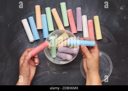 multicolored chalk in a plastic bucket on a black background, female hands fold objects into a container, top view Stock Photo