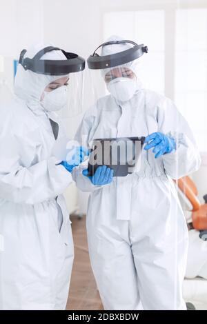 Stressed doctors dressed in ppe suit using tablet pc Stomatology team in dental office wearing protective suit agasint contagious coronavirus during global pandemic. Stock Photo