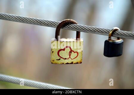 Old rusty yellow padlock with two hearts hanging on a metal fence next to small usual padlock outdoors Stock Photo
