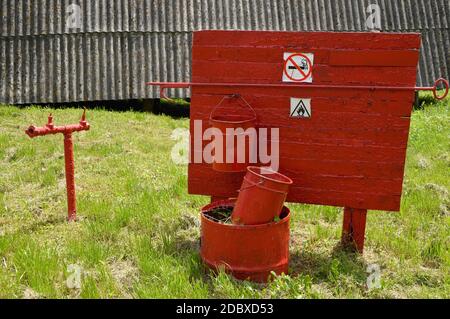Fire extinguishing equipment. Hydrant stand pipe and two buckets hanging on the board. Shabby and old with rusty red paint Stock Photo