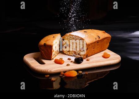 Sweet cupcake in the shape of bread. Cupcake with raisins, dried apricots, plums. Bread - cupcake on a wooden board on a black background, sprinkled w Stock Photo