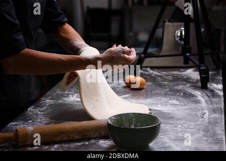 The chef prepares pastries in a professional kitchen. Knead the dough. Eggs, flour, rolling pin. Dark background. Stock Photo