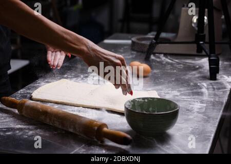 The chef prepares pastries in a professional kitchen. Knead the dough. Eggs, flour, rolling pin. Dark background. Stock Photo