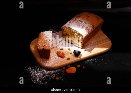 Sweet cupcake in the shape of bread. Cupcake with raisins, dried apricots, plums. Bread - cupcake on a wooden board on a black background, sprinkled w Stock Photo