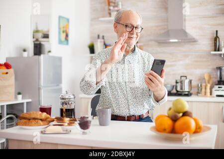 Elderly aged man waving during a video conference with family in kitchen. Elderly person using internet online chat technology video webcam making a video call connection camera communication conference call Stock Photo