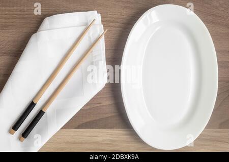 There is an empty white plate on the wooden table and wooden chopsticks on a napkin. The pattern of Japanese cuisine. The view from the top. Flat lay Stock Photo