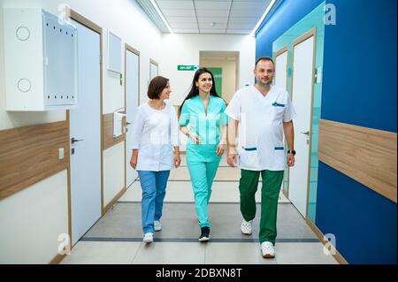 Three smiling doctors in uniform walking in clinic corridor. Professional medical specialist in hospital, laryngologist or otolaryngologist, gynecolog Stock Photo