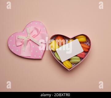 round baked multicolored macarons lie in a pink cardboard box in the shape of a heart on a beige background, top view Stock Photo