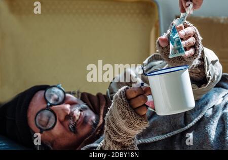 The depressed homeless man gets money help from passer-byes. he is unemployed and jobless due to the Coronavirus 19 pandemic. He lives by the sidewalk. Stock Photo