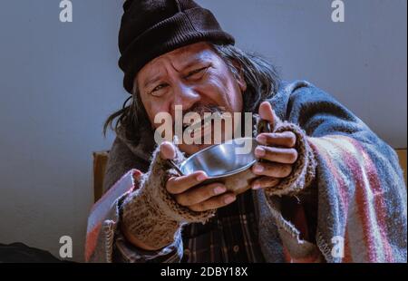 A pitiful homeless man with unhappy look asks for help by begging with bowl. Stock Photo