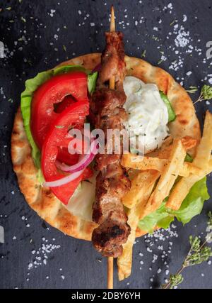 Souvlaki greek food. Grilled meat skewer served with fried potatoes, tomato and yogurt tzatziki on a pita bread, black stone background,  top view. Tu Stock Photo