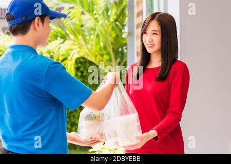 Asian young delivery man in blue uniform he making grocery service giving rice food boxes plastic bags to woman customer receiving front house under p Stock Photo