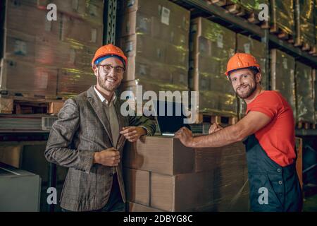Warehouse worker and manager standing near carboard boxes and looking positive Stock Photo