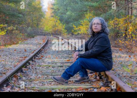 Pensive mature woman with grayish black hair and glasses, sitting on the train tracks, autumnal trees in the Meinweg nature reserve, old Iron Rhine Ra Stock Photo