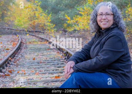Smiling mature woman with grayish black hair and glasses, sitting on train tracks, autumnal trees in the Meinweg nature reserve, old Iron Rhine railwa Stock Photo