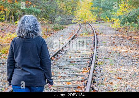 Mature woman walking among autumnal trees on the train tracks, grayish black hair, back to the camera, old Iron Rhine railway (IJzeren Rijn) in Meinwe Stock Photo