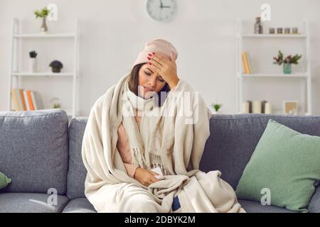 Young stressed woman in warm clothing and blanket sitting on sofa, touching forehead and feeling ill Stock Photo