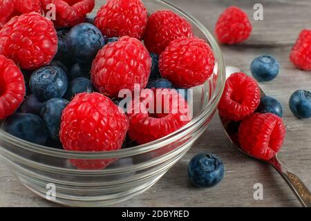 Small glass bowl of fresh raspberries and blueberries with spoon next to it, placed on gray wooden table. Stock Photo