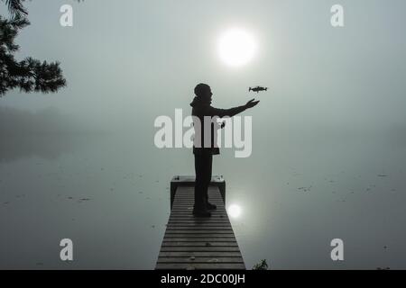 Man playing with the drone. Silhouette against the foggy landscape.Male operating the drone by remote control and having fun. Pilot flying drone. Use Stock Photo