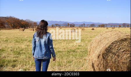 Young brunette woman in denim jacket standing near haystack in field with bales of hay. she looking at mountains and the autumn forest. Stock Photo