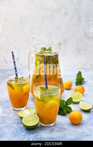 Homemade iced lemonade or tea with apricot, lime and mint. Fresh apricot mocktail in a jug and glasses on blue concrete background. Stock Photo