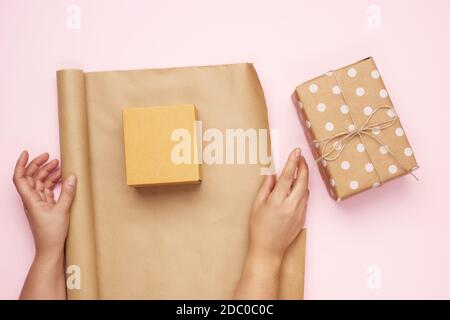 two female hands pack gifts in brown kraft paper on a pink background, top view Stock Photo