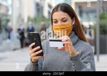 Portrait of young beautiful woman wearing protective mask chill out and make payment through credit card and smartphone app in the street Stock Photo