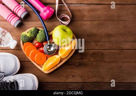 Top view of fresh fruits and vegetables in heart plate wood (apple, carrot, tomato, orange, broccoli) and sports equipment and doctor stethoscope on w Stock Photo