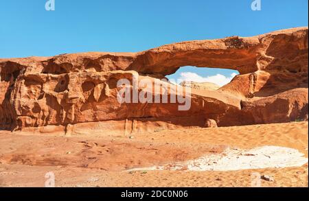 Little arc or small rock window formation in Wadi Rum desert, bright sun shines on red dust and rocks, blue sky above. Stock Photo