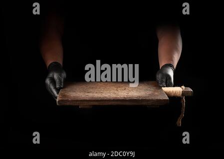 male hands in black latex gloves hold an empty vintage brown wooden chopping board, black background Stock Photo