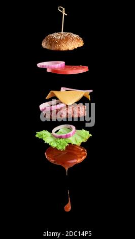 Flying ingredients of a classic cheeseburger: sesame bun, onion rings, tomato slices and a juicy barbecue cutlet on a black background. Fast food Stock Photo