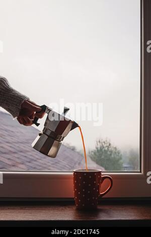 Woman pouring coffee from moka pot in the cup standing near the window Stock Photo