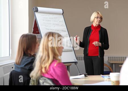 A middle-aged woman leads a management discussion - writing on a flip chart. Two younger women sit at a desk in the foreground. Stock Photo