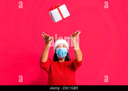 coronavirus christmas shopping concept. joyful adult woman with flying gifts box, face mask, santa claus outfit smiling on red background. christmas d Stock Photo