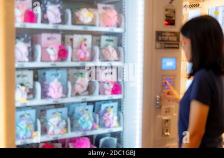 Blur Asian woman pressing button on vending machine to buy doll. Female tourists buying doll at automatic vending machine in airport shopping mall. Se Stock Photo