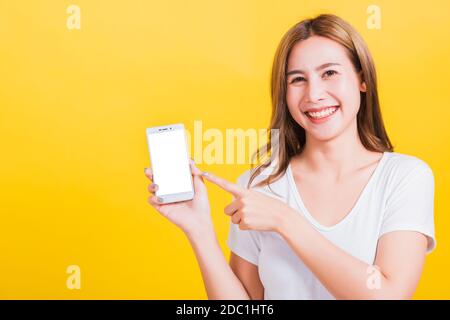 Asian Thai portrait happy beautiful young woman smile standing wear t-shirt making finger pointing on smartphone blank screen looking to camera isolat Stock Photo