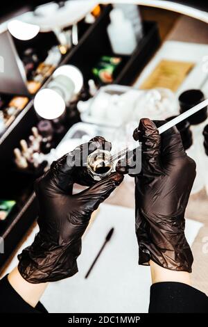 the hands of the master in black gloves hold a container with makeup liquid and a brush. Stock Photo