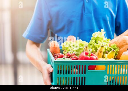 Asian grocery store delivery man wearing blue uniform and face mask protect he delivering fresh food vegetable in plastic box at door front home after Stock Photo