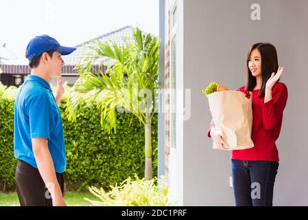Asian young delivery man in blue uniform making grocery service giving fresh vegetables and fruits and food in paper bag to woman customer at front ho Stock Photo