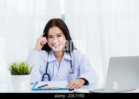 Asian doctor young beautiful woman smiling using working with a laptop computer and her writing something on paperwork or clipboard white paper at hos Stock Photo