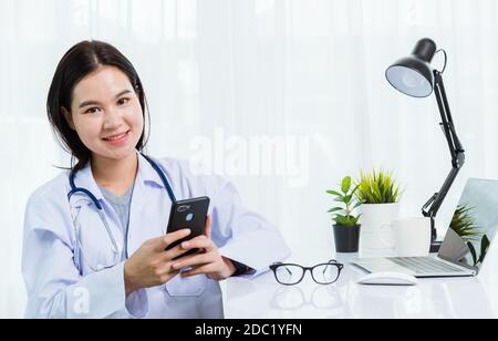 Asian doctor young beautiful woman smiling using working or holding with smart mobile phone and laptop computer at hospital desk office, technology he Stock Photo