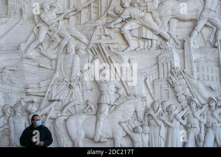 ROME ITALY  18 November 2020. A  man wearing a protective facemask stands in front of the large marble relief of Italian history depicting the former Fascist leader Benito Mussolini on a horse at the 1942 World fair held in Rome. The Italian government has imposed tighter lockdown restrictions from 6 November-3 December by closing museums, archeological sites to curb the spread of the soaring COVID-19 infection rates. Credit: amer ghazzal/Alamy Live News Stock Photo