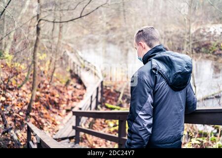 Man wearing a corona virus mask while walking outdoors in the nature in winter or autumn. Young person hiking the forest during coronavirus pandemic. Stock Photo