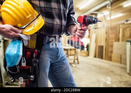 Carpenter in the carpentry workshop, holding a helmet, goggles, leather gloves, surgical mask to prevent infection by the coronavirus and cordless dri Stock Photo