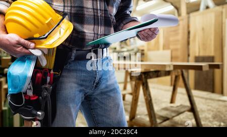 Carpenter in the carpentry workshop, holding a helmet, goggles, leather gloves, surgical mask to prevent infection by the coronavirus and notebook for Stock Photo