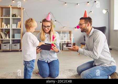 Happy parents give a gift and a cake to their son's birthday in a cozy living room. Stock Photo